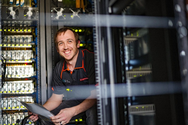 A smiling male stands in the BAE Systems Australia server room, surrounded by rows of servers and blinking lights.